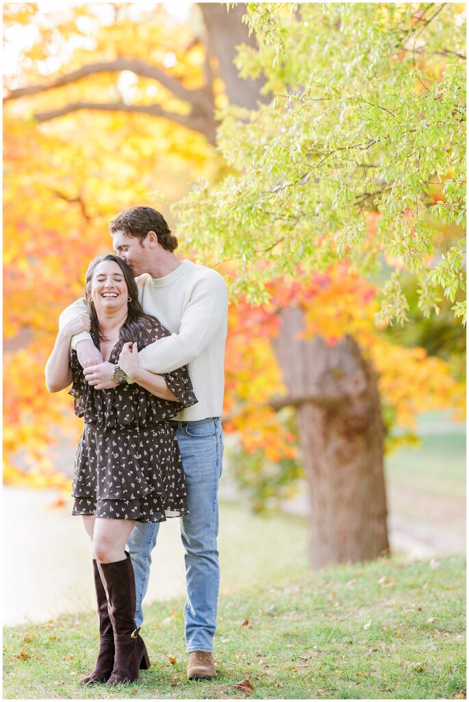 The couple smiles and embraces on a quiet sidewalk at Bartlet Mall in Newburyport, with red ivy on a stone wall and yellow-leaved trees in the background.