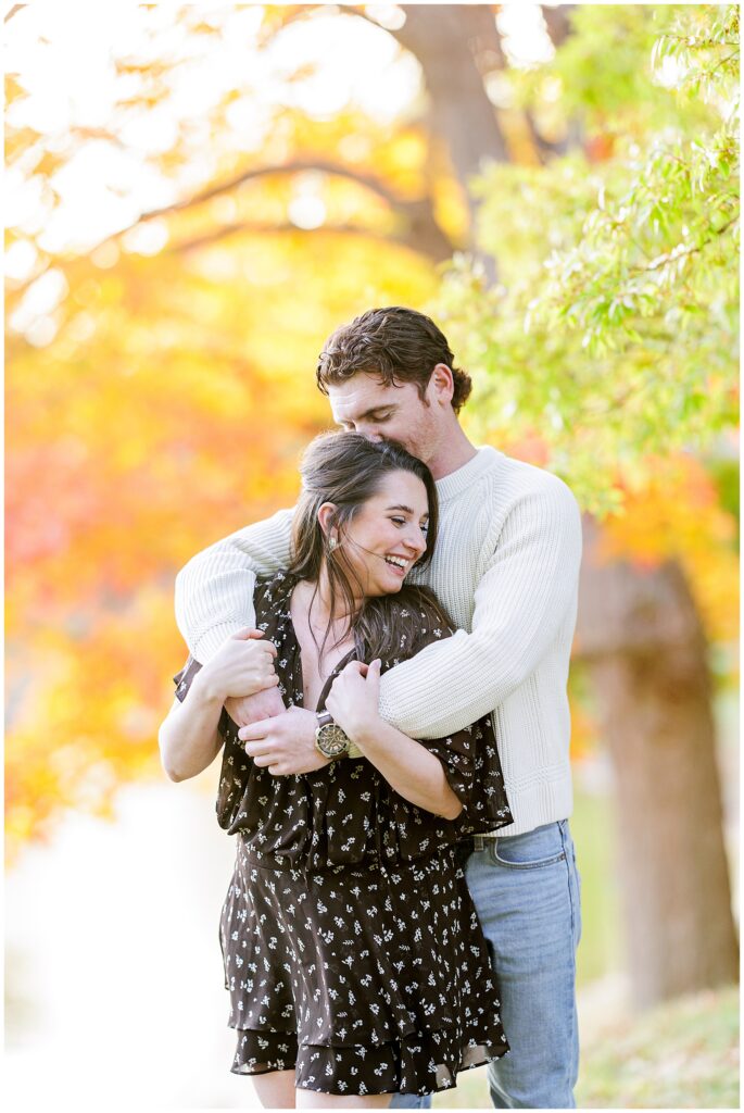 At Bartlet Mall in Newburyport, the couple stands in front of a pond and vibrant fall trees, laughing together as the man kisses the woman’s head.
