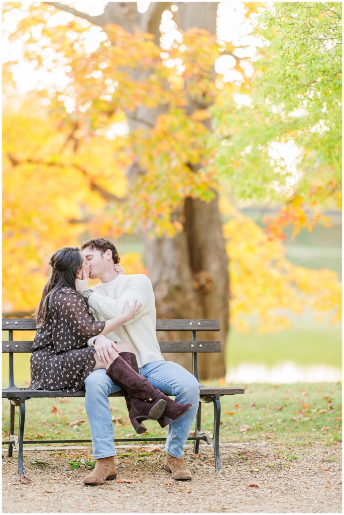 The couple shares a kiss while sitting on a bench by the water at Bartlet Mall in Newburyport, framed by trees in full autumn color.
