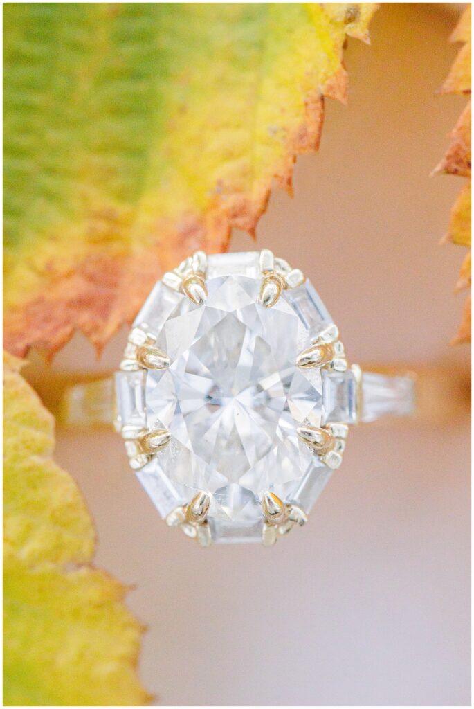 Close-up of an oval-cut diamond engagement ring with baguette side stones, displayed on a yellow autumn leaf at Bartlet Mall in Newburyport.