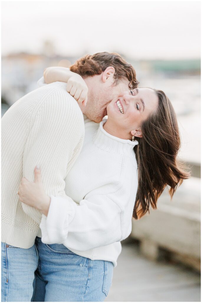The couple laughs together as the man kisses the woman’s neck during their engagement session along the boardwalk at the Newburyport waterfront.