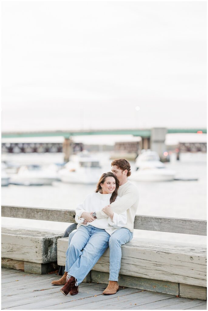 The couple sits on a wooden bench along the Newburyport waterfront, with boats and the bridge in the background during their fall engagement shoot.
