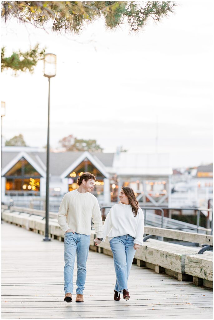 The couple walks hand-in-hand down the wooden boardwalk at the Newburyport waterfront, surrounded by warm glow from nearby buildings in the background.