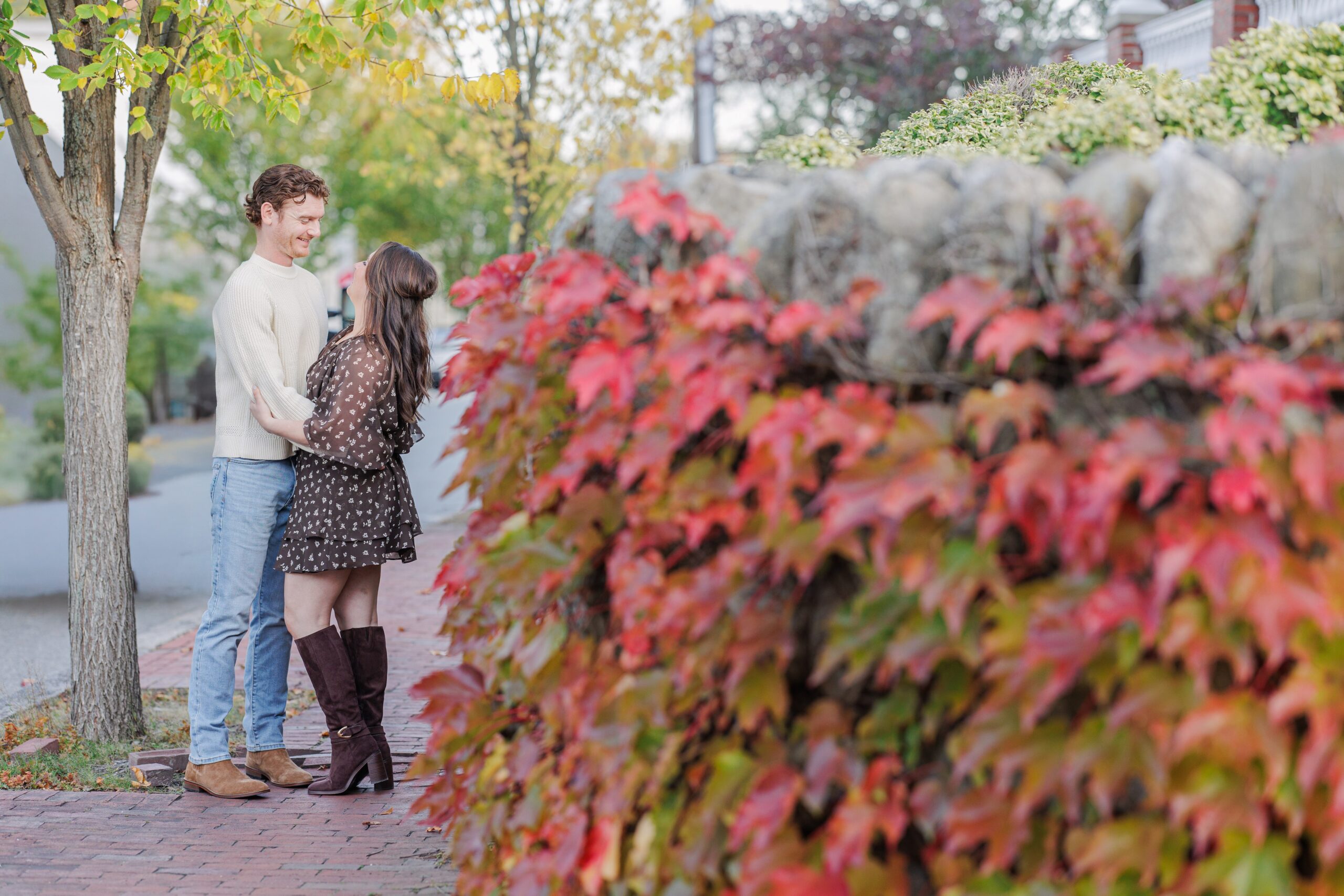 The couple stands on a red brick sidewalk next to a stone wall covered in vibrant red ivy at Bartlet Mall in Newburyport, surrounded by early fall foliage.
