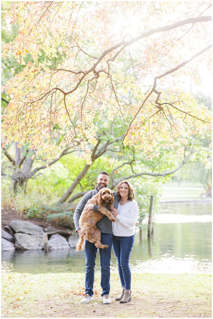 A couple stands smiling under colorful autumn branches, holding a fluffy brown dog in front of a pond and rocks.