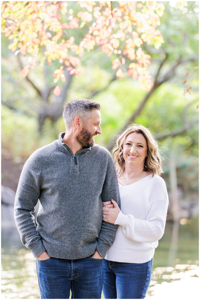A close-up of the couple standing arm-in-arm, smiling at each other, surrounded by soft fall foliage.
