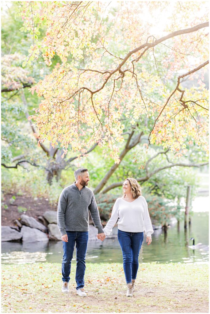 The couple walks hand-in-hand near the pond, beneath glowing yellow and pink fall leaves.
