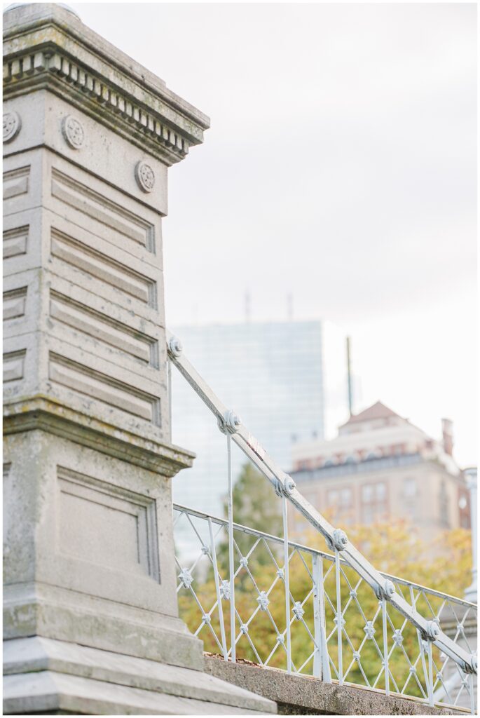 A stone post and metal railings of the Boston Public Garden footbridge, with city buildings in the background.
