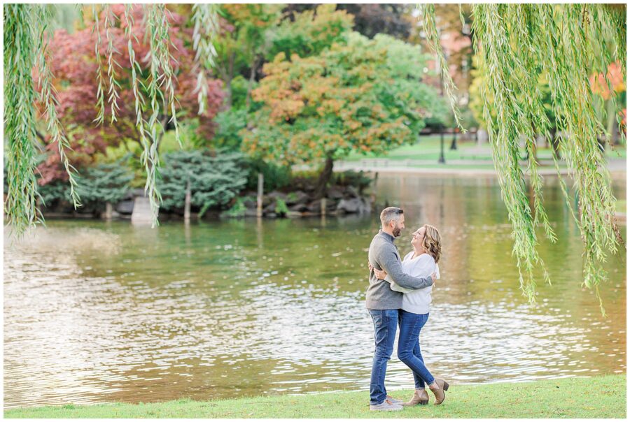 The couple embraces near the pond, framed by hanging willow branches and vibrant autumn trees across the water.