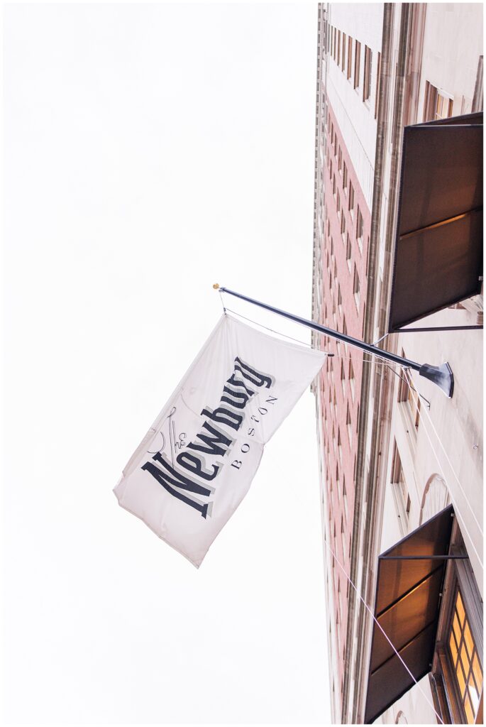 A white flag with “The Newbury Boston” in bold black lettering waves on a flagpole attached to the side of the hotel building, shot from below against an overcast sky.