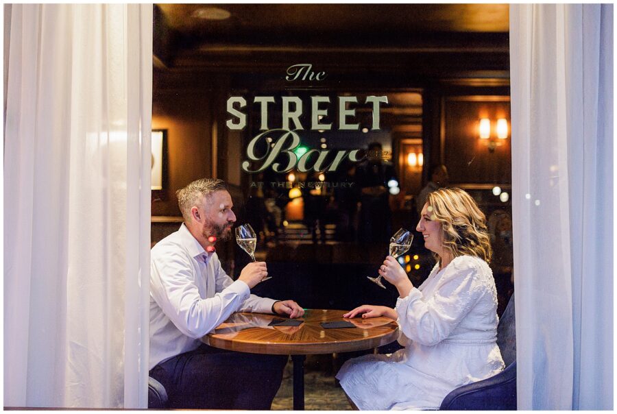 A couple sits inside The Street Bar at The Newbury Boston, smiling at each other and toasting with wine glasses, viewed through the window framed by white curtains.