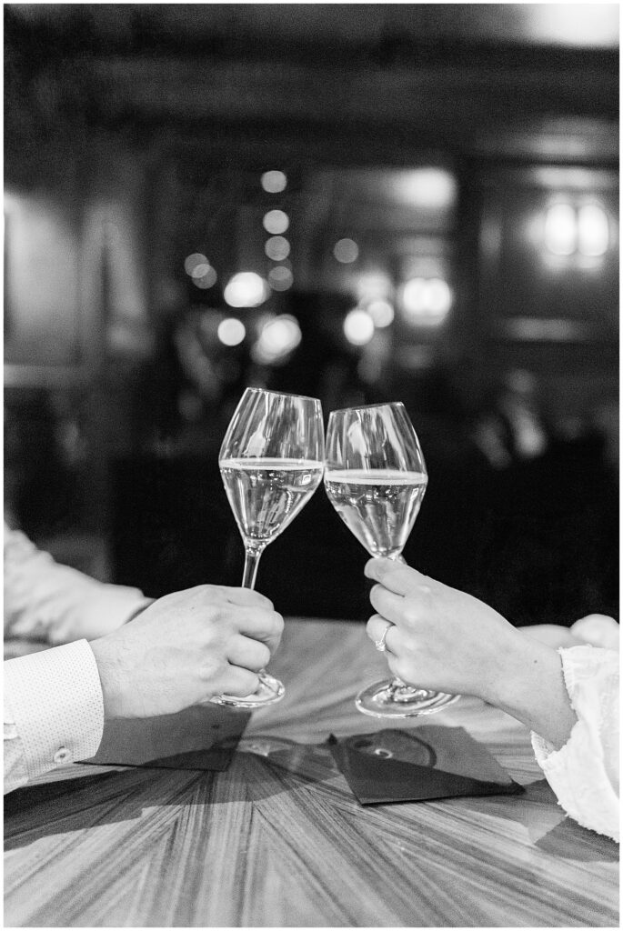 Close-up black-and-white image of two hands clinking wine glasses over a wood table at The Street Bar in The Newbury Boston.