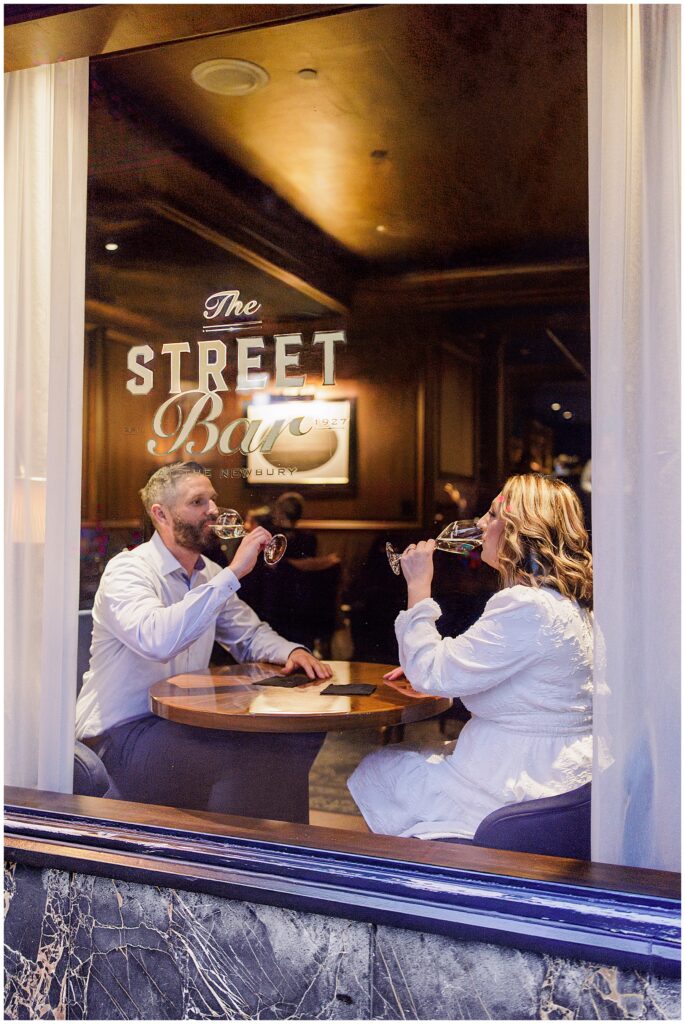 The couple clinks glasses while seated at a round table inside The Street Bar at The Newbury Boston, photographed from outside the window with the bar’s name visible on the glass.