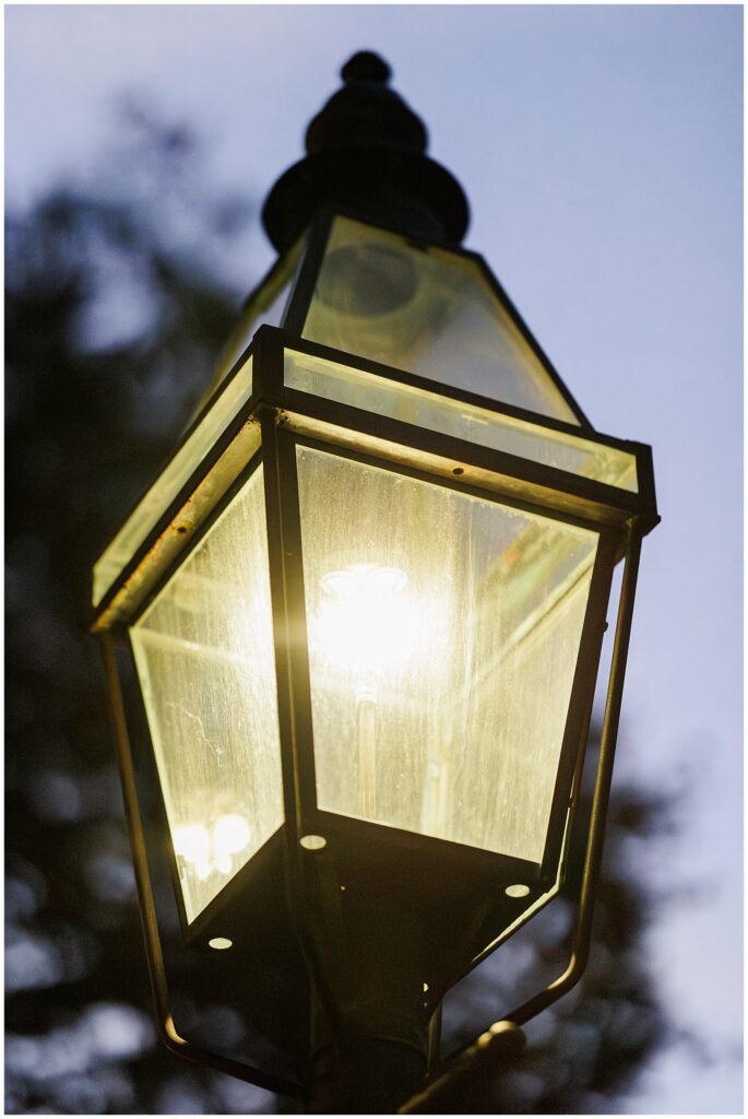 A close-up of a glowing vintage-style streetlamp at dusk in Beacon Hill, Boston, with soft focus trees in the background.