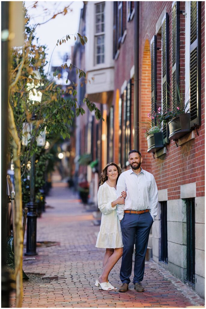 A couple stands arm in arm on a cobblestone sidewalk in Beacon Hill, Boston, in front of a red brick building with black shutters and flower boxes.