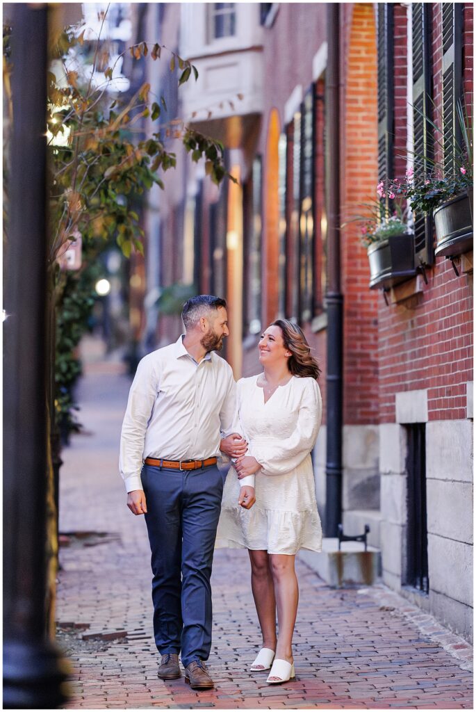 The couple walks closely together on a brick sidewalk in Beacon Hill, Boston, smiling at each other, surrounded by warm evening light and classic row houses.