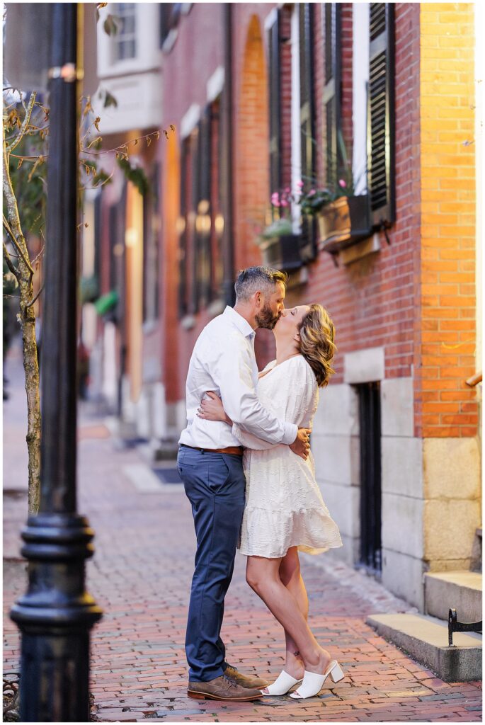 The couple kisses on a charming brick sidewalk in Beacon Hill, Boston, with warm light reflecting off the red brick buildings and classic black lamp posts lining the street.