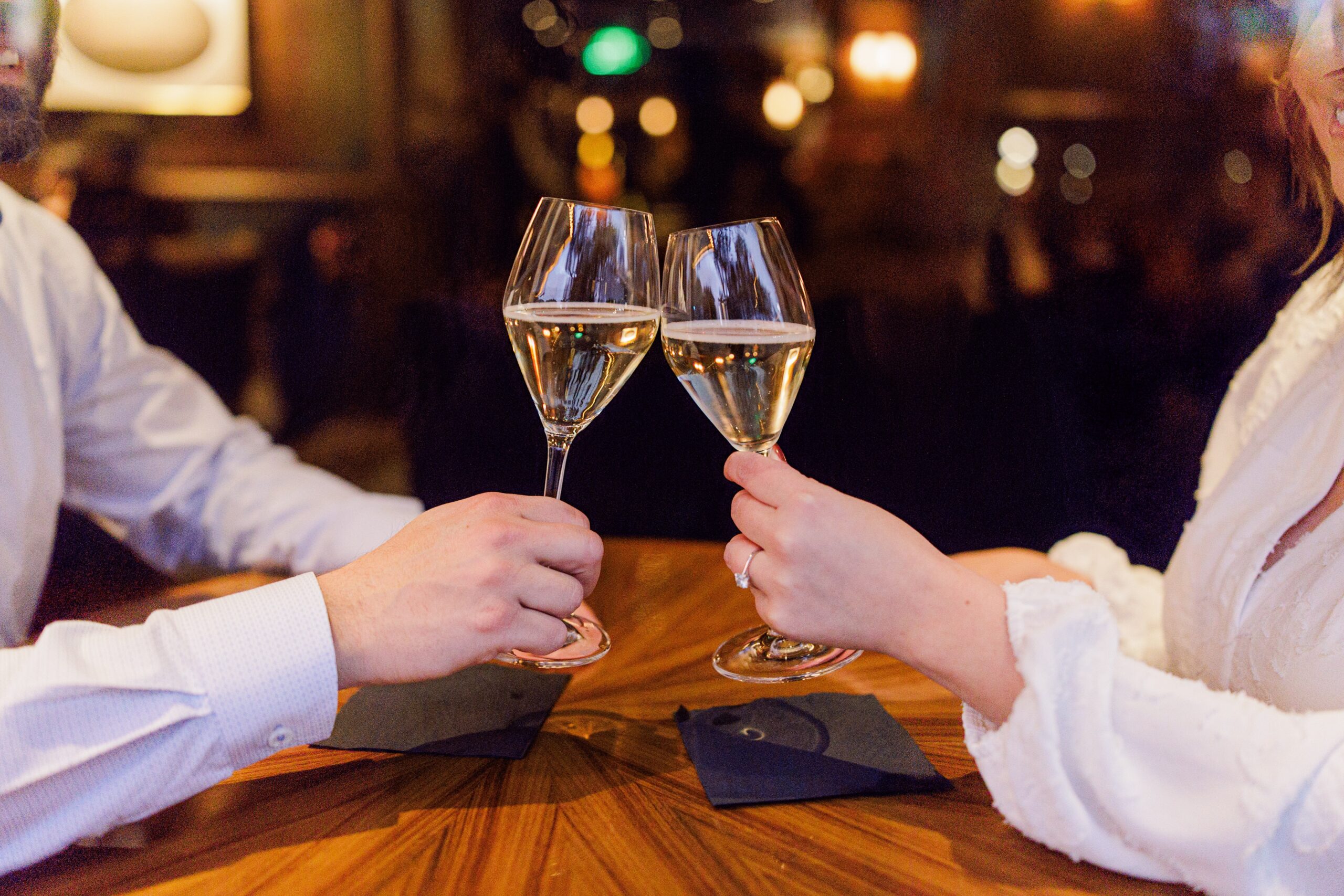 A close-up of a toast with two glasses of white wine inside The Street Bar at The Newbury Boston, with warm lighting and soft background bokeh.