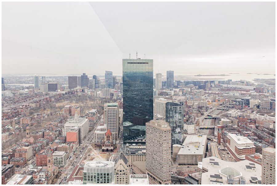 A panoramic view of downtown Boston taken from View Boston at the top of the Prudential Center, showing the John Hancock Tower centered among other skyscrapers, with Boston Harbor in the distance under an overcast sky.