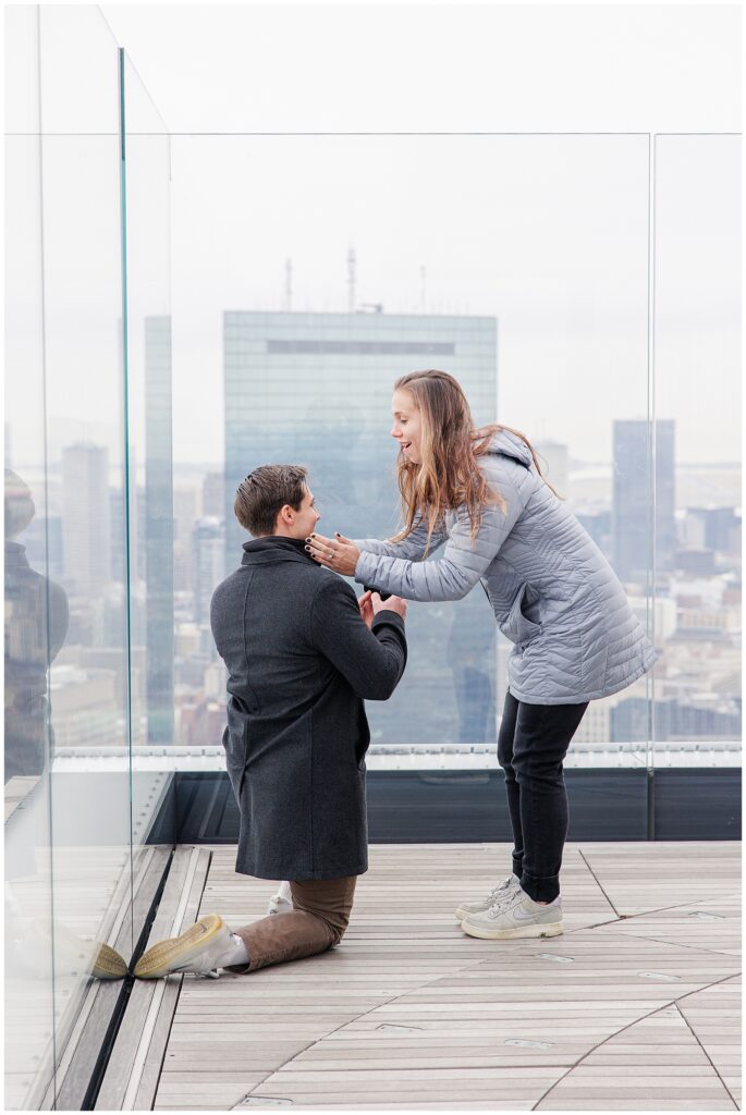 The woman leans forward, smiling with joy, placing her hands on the shoulders of the man who remains on one knee, moments after the proposal.