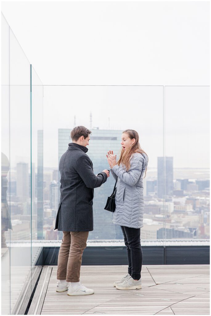 The couple stands facing each other on the observatory deck as the man places the engagement ring on the woman’s finger; she appears surprised and excited.