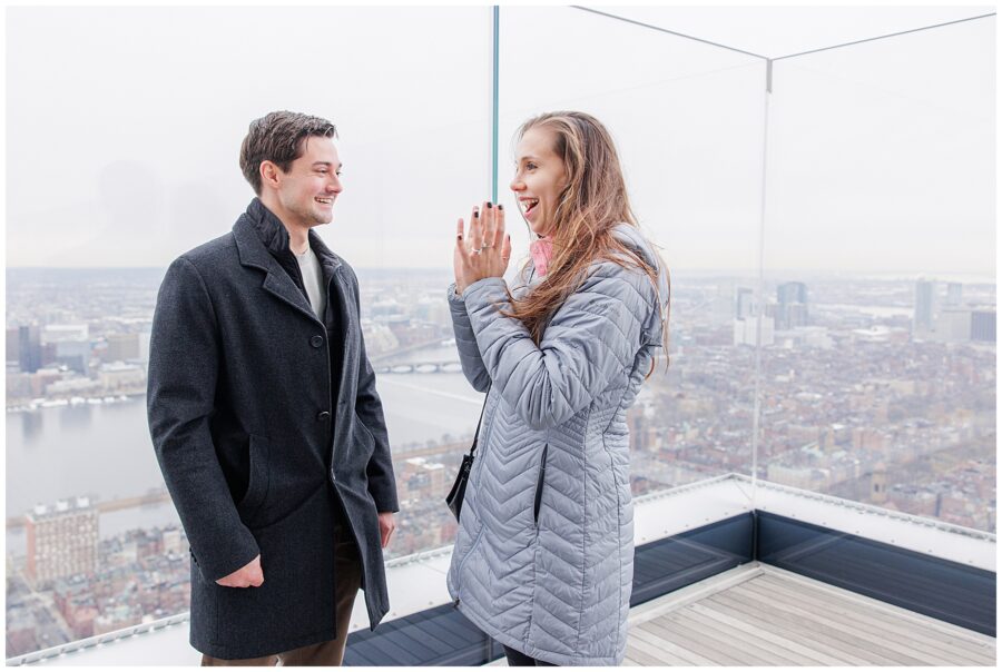 The couple smiles at each other, standing close together on the rooftop deck with the Charles River and cityscape visible behind them.