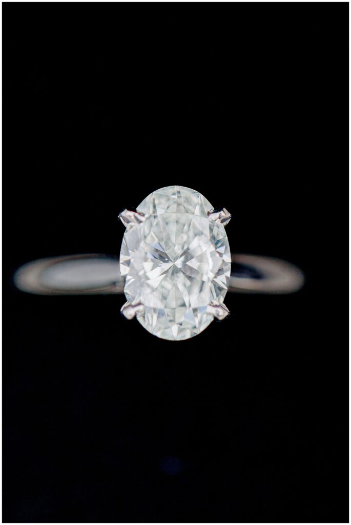 A close-up shot of a sparkling oval-cut diamond engagement ring in a four-prong setting, photographed against a black background.