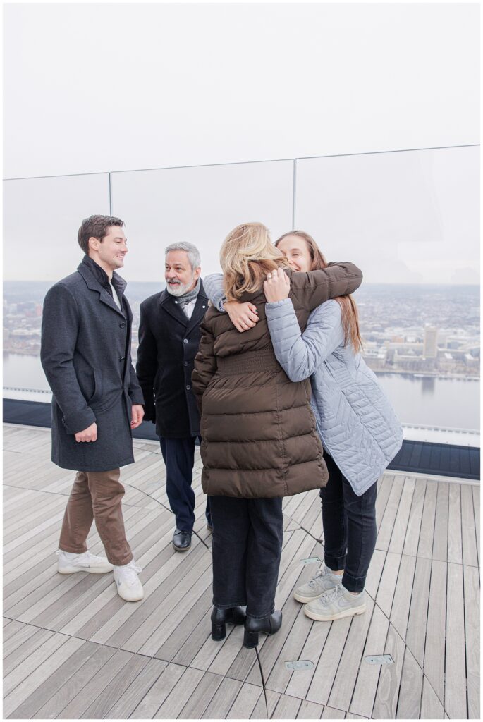 A newly engaged woman hugs an older woman while two men smile nearby on the rooftop observatory at View Boston, overlooking the city.