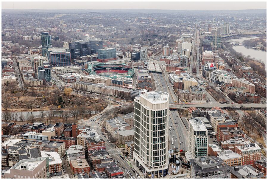 A panoramic cityscape featuring Fenway Park, the CITGO sign, and surrounding Boston neighborhoods, taken from above on an overcast day.