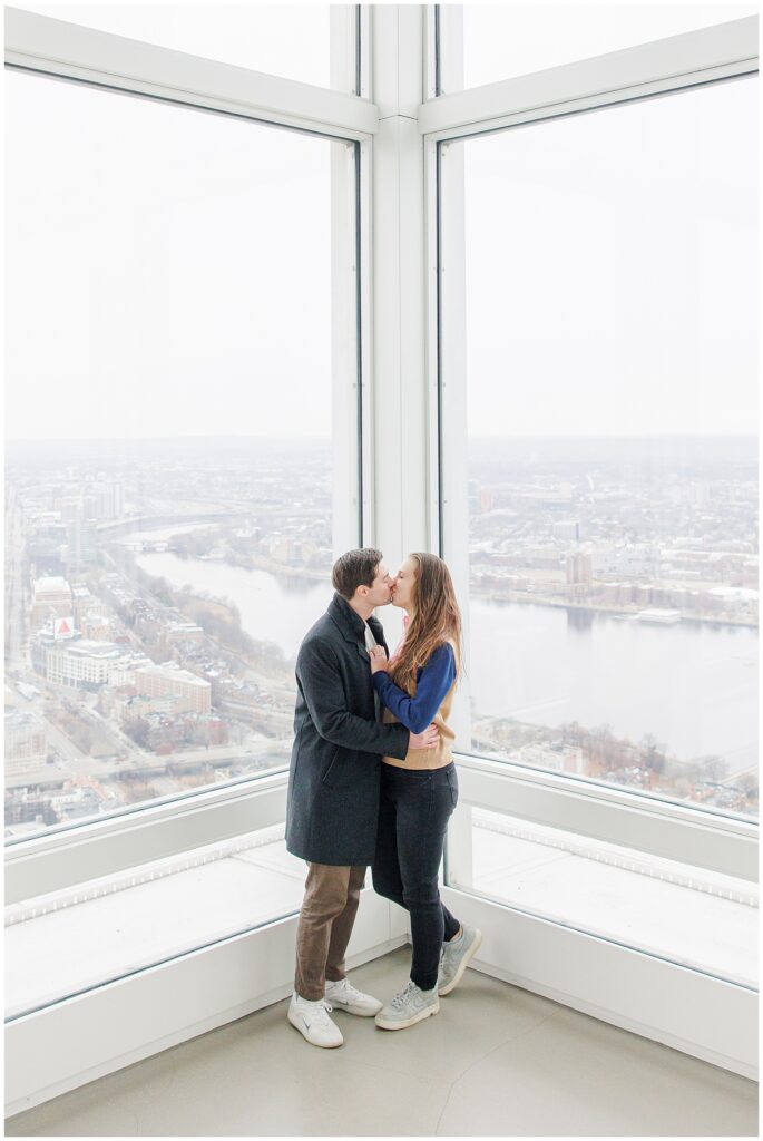 A couple kisses while standing in the corner of a glass-walled observation area at View Boston, with the Charles River and Boston skyline behind them.