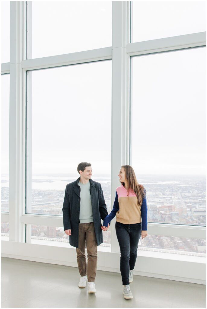 A couple holds hands and smiles at each other while walking inside the View Boston observatory, surrounded by tall windows with panoramic views of the city.