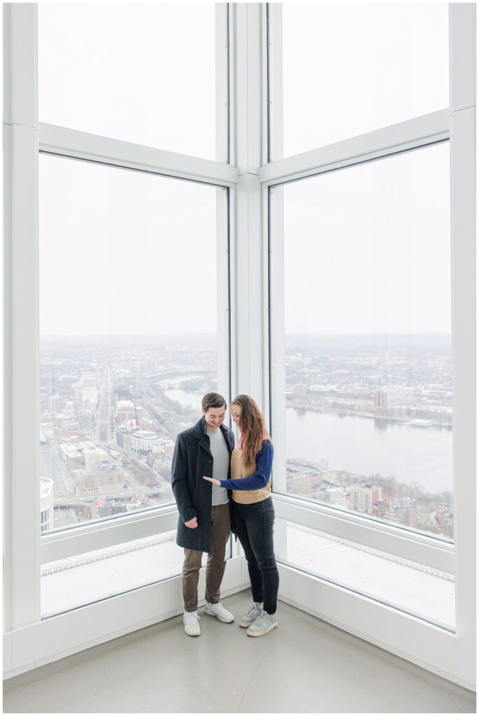 A couple stands together by a corner window at View Boston, looking down at the woman’s hand as she shows off her new engagement ring.