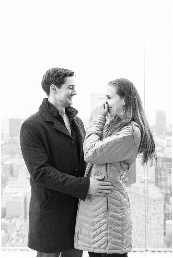  A black-and-white photo of the couple smiling and standing close together at View Boston, with the woman covering her mouth and the city skyline faintly visible behind them.