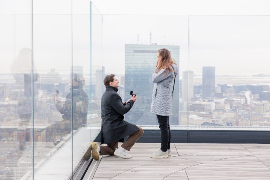 A man kneels and holds out a ring box while proposing to a woman on the glass-walled outdoor deck at View Boston, with reflections visible on the panels and the John Hancock Tower behind them.