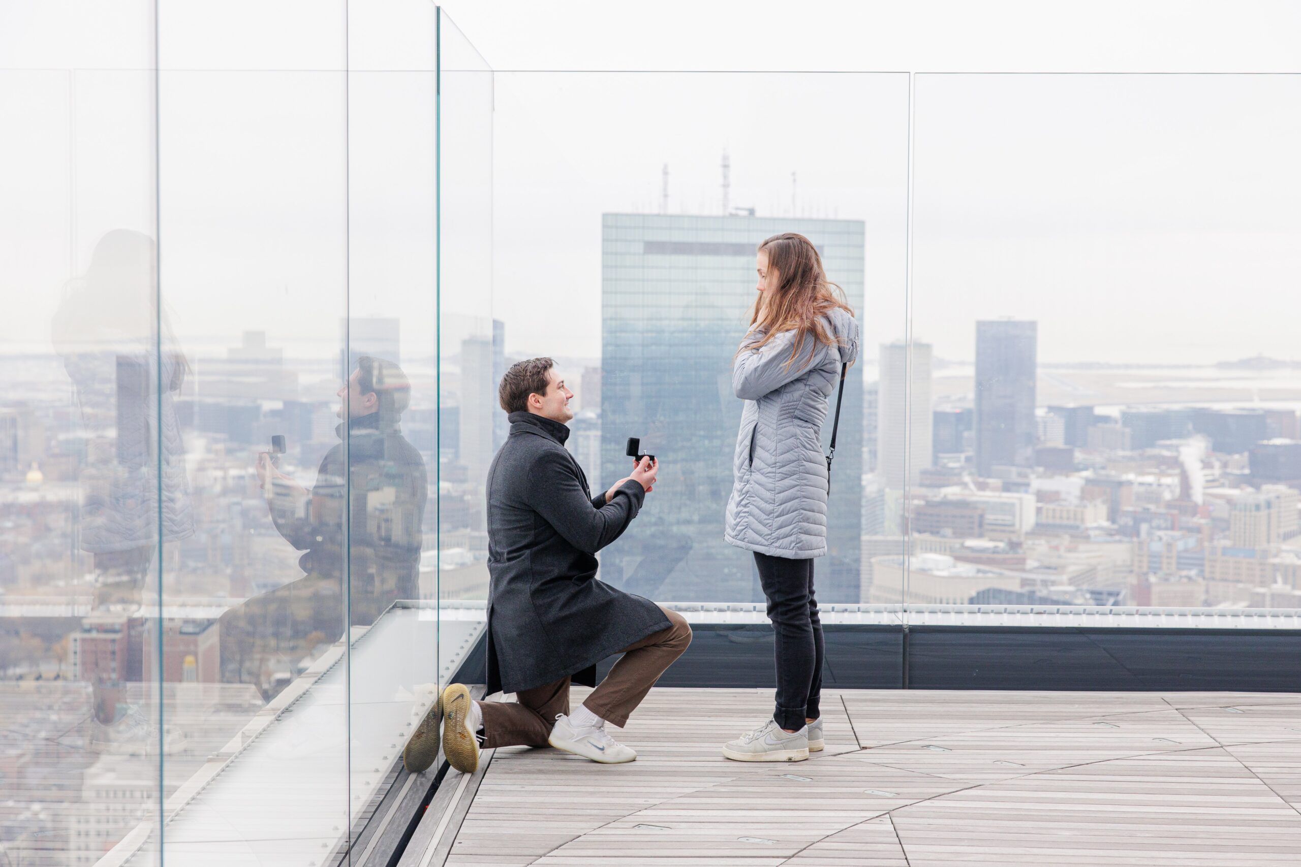 A man kneels and holds out a ring box while proposing to a woman on the glass-walled outdoor deck at View Boston, with reflections visible on the panels and the John Hancock Tower behind them.