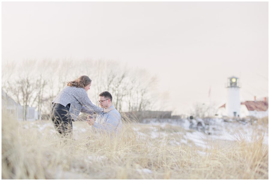Man getting down on one knee in the dunes for a Chatham Lighthouse Beach proposal with the lighthouse softly blurred in the background at Chatham Lighthouse Beach in Chatham, MA Cape Cod.