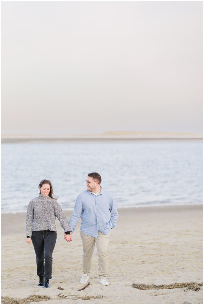 Couple holding hands and walking along the shoreline during their Chatham Lighthouse Beach proposal at Chatham Lighthouse Beach in Chatham, MA Cape Cod.