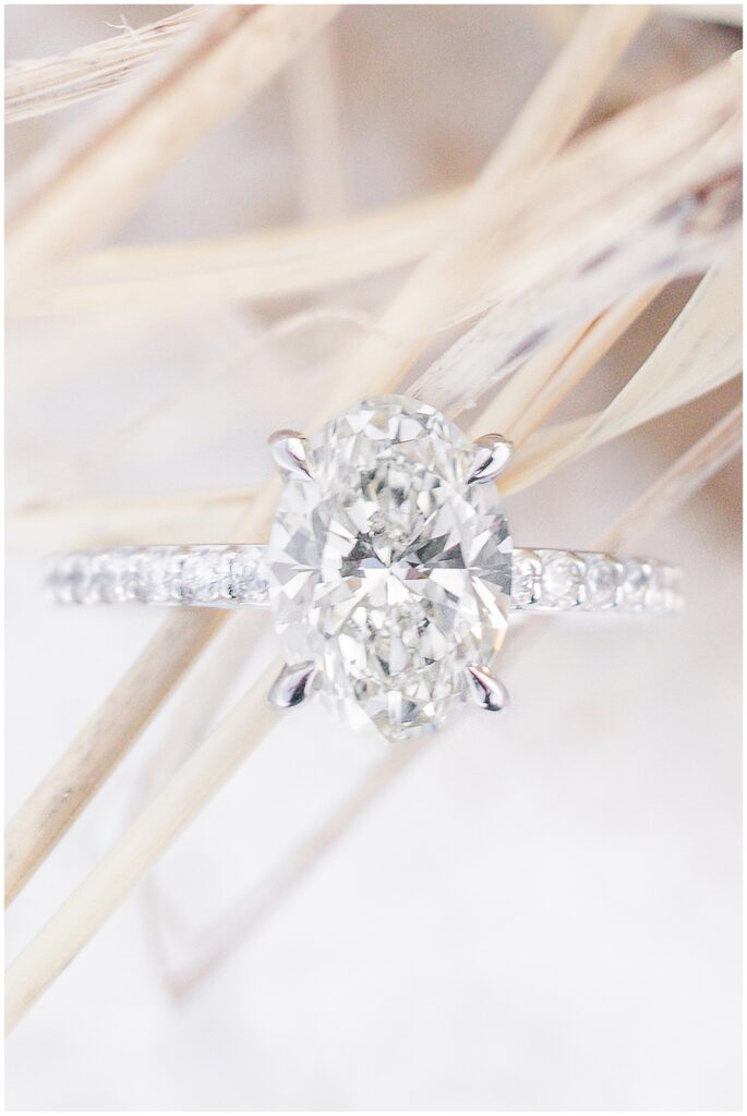 Close-up of oval diamond engagement ring photographed in soft beach grass after a Chatham Lighthouse Beach proposal in Chatham, MA Cape Cod.