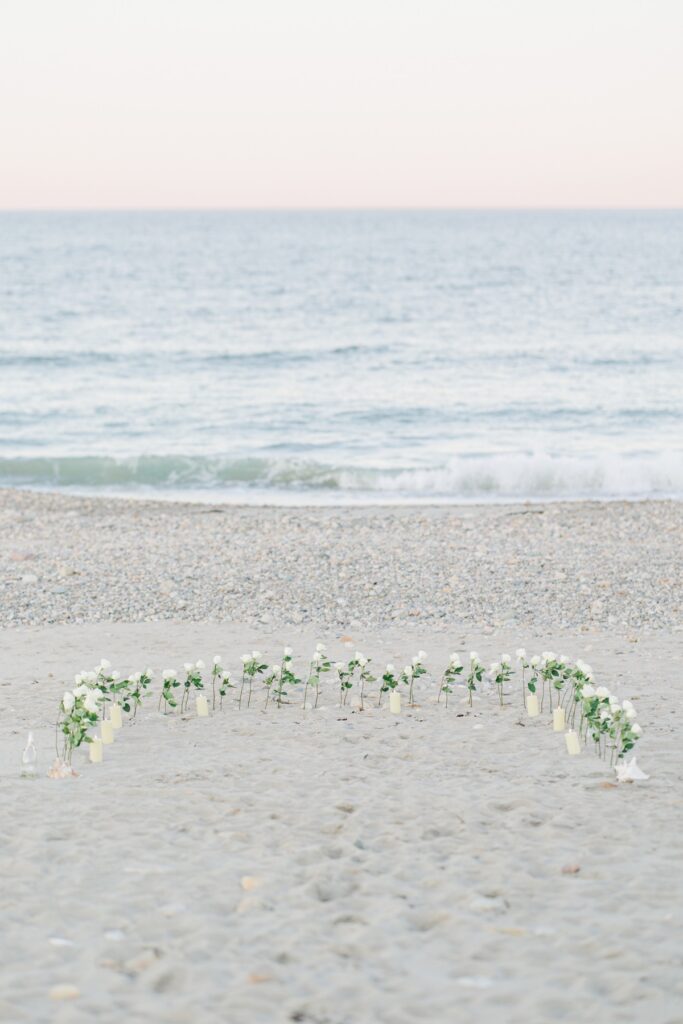 Horseshoe of white roses arranged in the sand for a romantic Duxbury Beach proposal overlooking the ocean