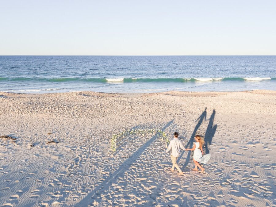 Couple walking toward a rose-lined setup on Duxbury Beach before their proposal at sunset