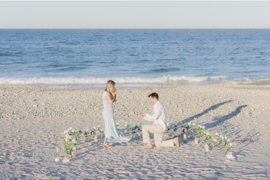 Man proposing on one knee surrounded by white roses during a Duxbury Beach proposal