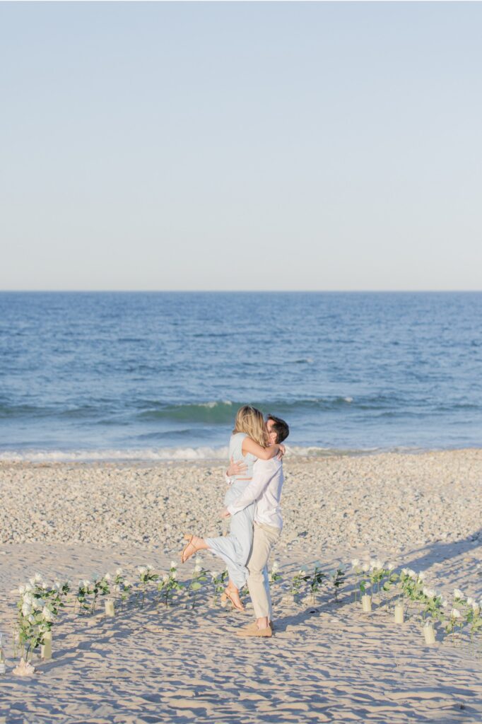 Couple kissing on the beach surrounded by white roses after a romantic proposal