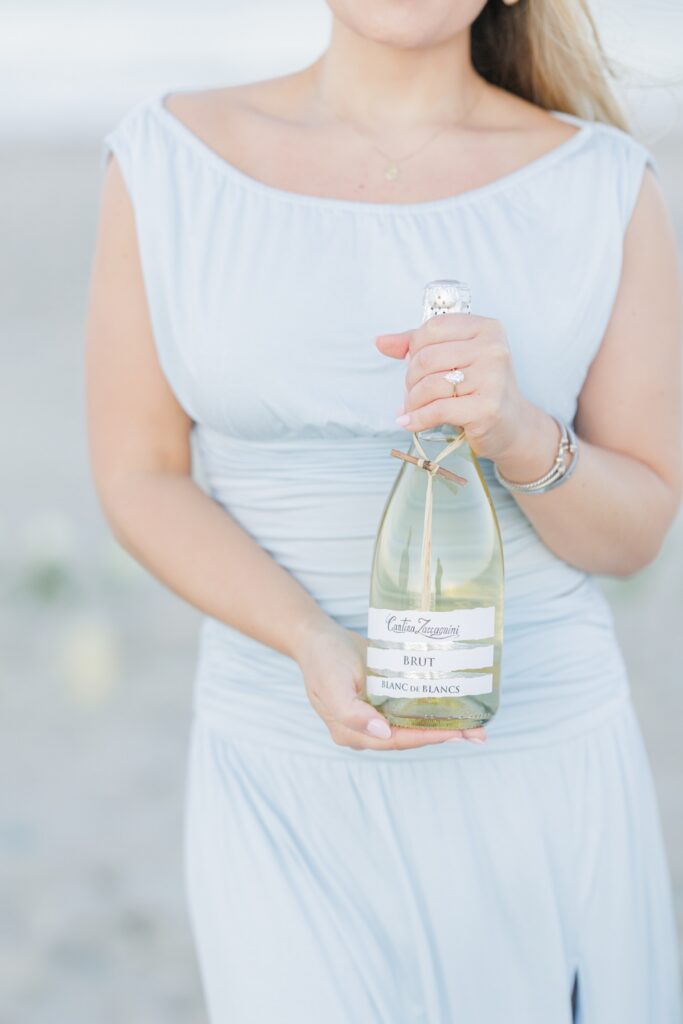 Bride holding champagne bottle and showing engagement ring after beach proposal