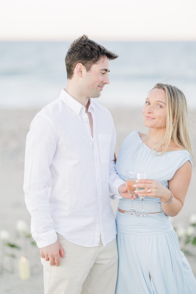 Engaged couple holding champagne glasses and smiling on Duxbury Beach after proposal