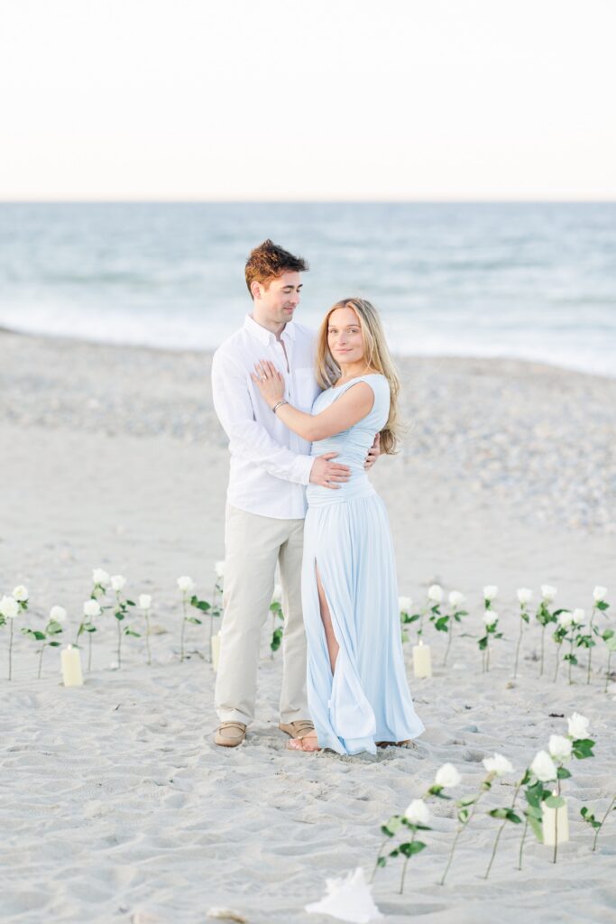 Engaged couple posing on the beach surrounded by white roses after Duxbury Beach proposal