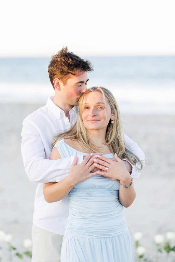 Close up of groom kissing bride’s head showing engagement ring during beach proposal session