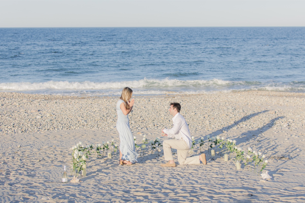 Man proposing on one knee surrounded by white roses on Duxbury Beach