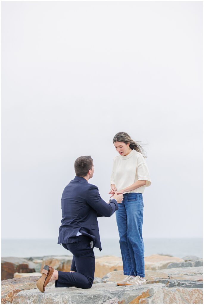 Scituate Lighthouse proposal ring placement moment with surprised bride to be on coastal rocks