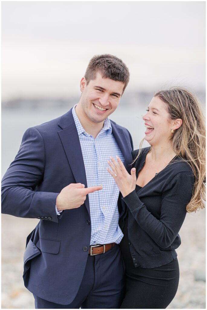 Playful couple showing off engagement ring after Scituate Lighthouse proposal with coastal backdrop