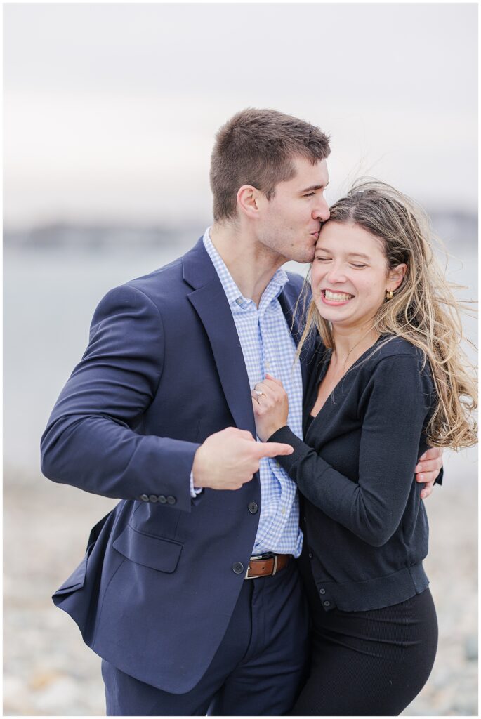 Groom kissing bride to be’s forehead while pointing to engagement ring after Scituate Lighthouse proposal